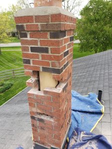 Chimney Repair 8 A brick chimney on a roof with a section of bricks removed, revealing a wooden post inside. Blue moving blankets are spread on the shingles nearby, and trees and a street are visible in the background.