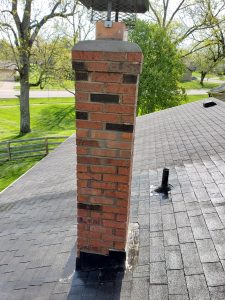 Chimney Repair 7 A brick chimney on a shingled roof is visibly leaning to one side, with metal flashing at its base. Trees and a grassy yard are visible in the background, along with a street and parked cars.