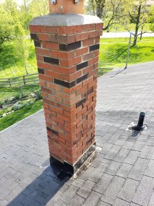 Chimney Repair 6 A brick chimney rises from a shingled roof, surrounded by green trees, a fence, and a grassy yard on a sunny day.