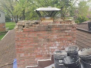 A damaged brick chimney with missing and crumbling bricks at the top, a metal chimney cap, and several black buckets placed on the nearby roof. Green trees and houses are visible in the background.