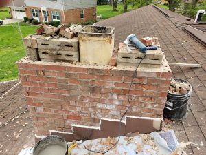 Chimney Crowns Repair 19 A brick chimney on a roof, partially disassembled, with bricks, mortar, a bucket, and tools scattered on top and around it. The chimney shows signs of ongoing repairs or construction work.