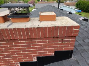 Chimney Crowns Repair 20 A close-up view of a brick chimney on a shingled roof, showing cracks in the top cement cap and some damaged or missing mortar between the bricks.