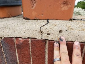 A close-up of a persons hand touching a cracked concrete cap on top of a brick chimney, with visible cracks near the base of the clay chimney flue. The hand has paint stains and a silver ring.