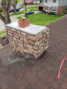 Chimney Crowns Repair 23 A stone chimney with a concrete cap and brick flue sits on a shingled roof. A red strap lies nearby, and suburban houses, trees, and a U.S. flag are visible in the background.