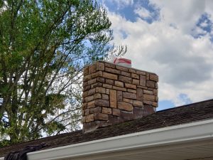 Chimney Crowns Repair 25 A brick chimney on a house roof with a red-and-white bag placed on top, set against a partly cloudy sky and green leafy trees in the background.