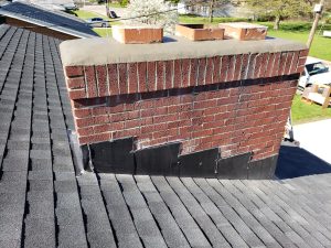 Chimney Crowns Repair 26 A red brick chimney on a shingled roof with black metal flashing along the base, extending up the side of the chimney. Nearby houses, grass, and a blooming tree are visible in the background.