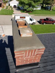 A brick chimney with a fresh layer of cement on top and three capped flues, viewed from above. Two trucks and a lawn are visible on the street behind the house.