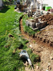 A curved trench is dug along the edge of a grassy yard. A power tool and yellow extension cord lie nearby. Stacks of concrete blocks and dirt piles are visible next to a house and deck in the background.