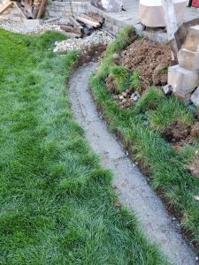 A curved trench dug along the edge of a grassy lawn next to a stacked stone wall, with piles of dirt and construction materials nearby.