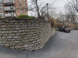 A stone retaining wall lines the edge of a paved parking lot near an apartment building, with a pickup truck parked by the curb and bare trees in the background on a cloudy day.