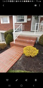 Brick And Stone Repairs 11 Brick repair of Red brick steps and a walkway lead up to a brick house with white railings and shutters. Bushes and a yellow shrub border the walkway, and a window with white trim is visible above the entrance.
