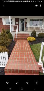 A brick walkway leads to a set of stairs and the front door of a house with a porch, white railings, trimmed bushes, a white picket fence, and a wreath on the door.