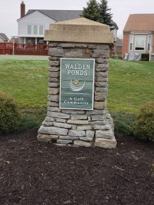 A stone sign in front of green grass reads Walden Ponds: A Golf Community, with houses visible in the background on an overcast day.