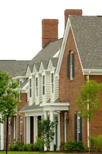 Chimney Repair 4 Row of two-story suburban houses with brick and light-colored siding, multiple chimneys, gabled roofs, white columns, green shutters, and landscaping with bushes and small trees in front.