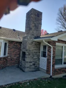 Chimney Repair 5 A brick house with a stone chimney on a small concrete patio. The chimney is tall and rectangular, built into the exterior wall between windows and a white door, with leaves scattered on the grass.